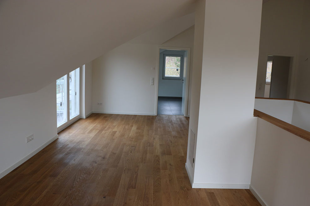 Zimmer mit Holzboden und weißen Wänden, in der Mitte des Raumes führt eine Treppe in das Erdgeschoss