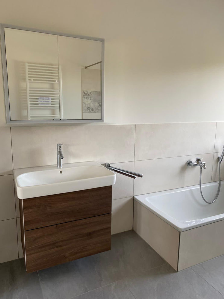Badezimmer mit hellen Wandfließen, einem Waschbecken mit Spiegelschrank, eine Badewanne und einem graue Fließenboden